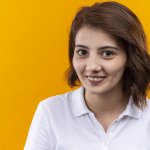 young girl with short hair wearing polo shirt looking at camera with smile on face standing over orange background